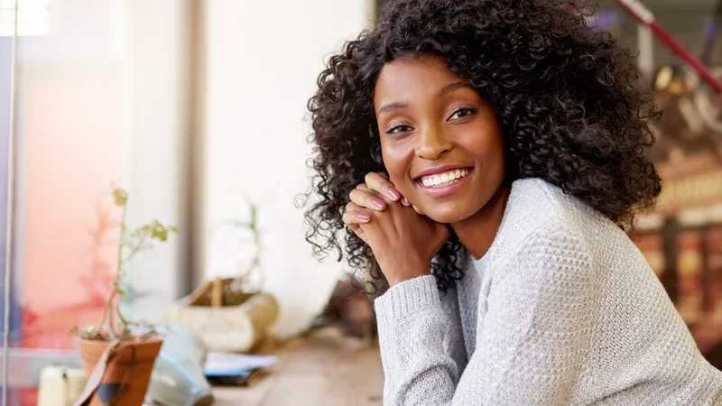 Black woman with long curly hair, in gray sweater, sitting at kitchen table.