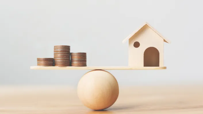 A picture of wooden scale that has pennies on one side and a house on the other.