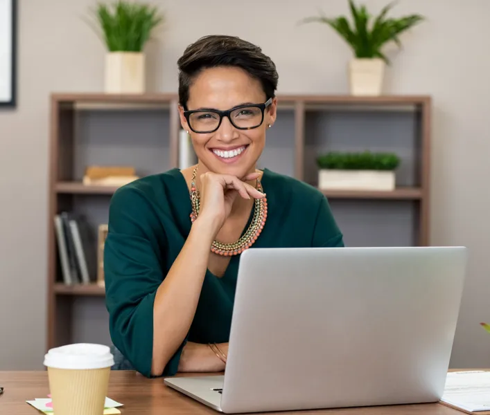 Diverse woman with glasses smiling sitting at a desk in front of laptop