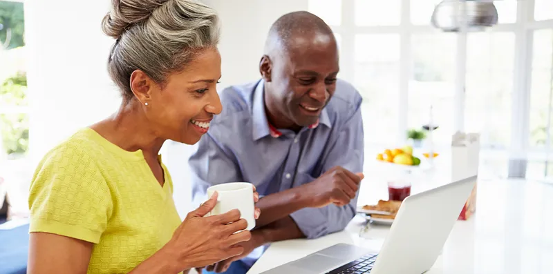 couple holding coffee at kitchen table smiling, looking at laptop