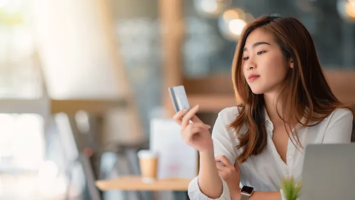 woman with long hair looking at credit card in hand at coffee shop table with laptop