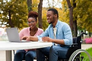 two smiling people outside at table looking at laptop, one sitting in chair, one person sitting in wheelchair