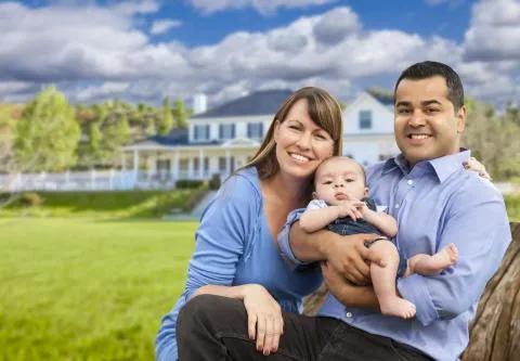 A young family holding a baby sitting in front of their white house with dark gray shutters.