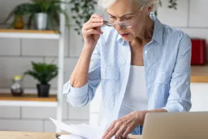 An elderly person looking through paperwork while her laptop is open.