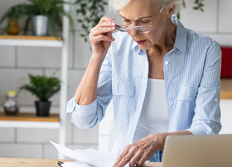 An elderly person looking through paperwork while her laptop is open.