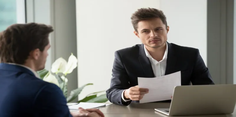 Two men discuss paperwork at a desk.