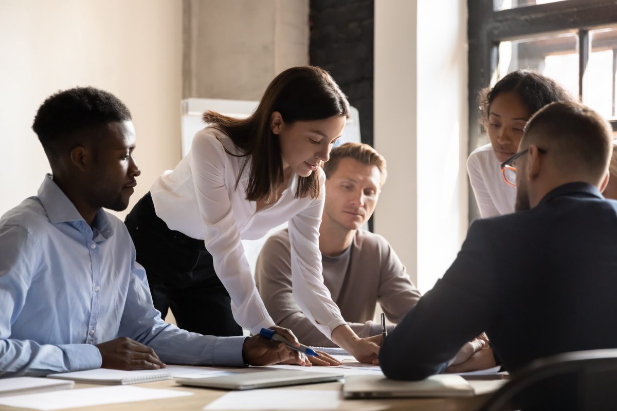 A group of people in a meeting room with a woman leading a discussion.